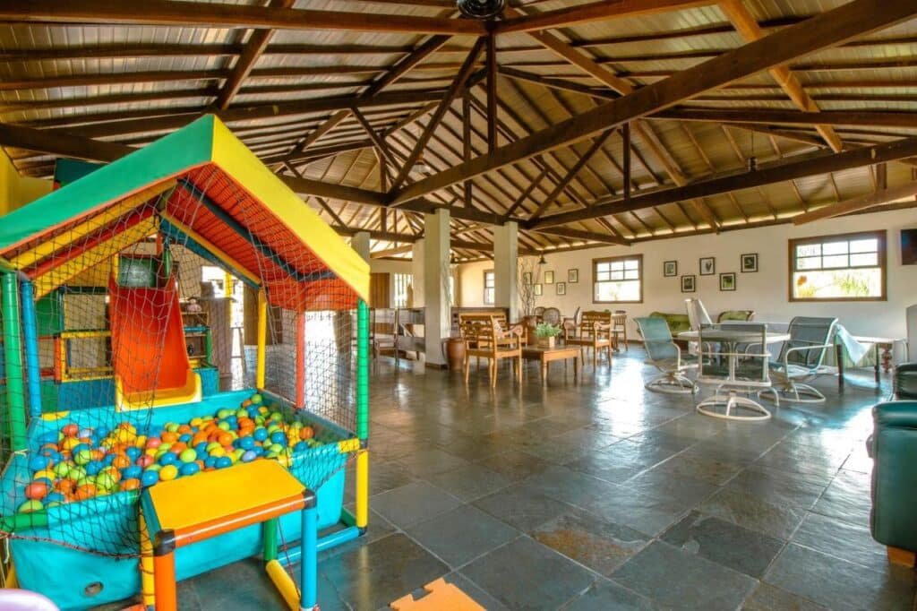 A imagem mostra um amplo salão de jogos e convivência com piso de pedra escura e teto alto com vigas de madeira expostas. À esquerda, há uma estrutura colorida de brinquedos infantis, incluindo uma piscina de bolinhas e um pequeno escorregador laranja. No restante do salão, há mesas com cadeiras de madeira e metal, além de sofás e poltronas, criando um espaço confortável para socialização. As paredes são brancas e decoradas com quadros, e grandes janelas permitem a entrada de luz natural, iluminando o ambiente de forma suave e agradável. Esse é o Hotel Fazenda Minas Real, um dos hotéis-fazenda em Minas Gerais.