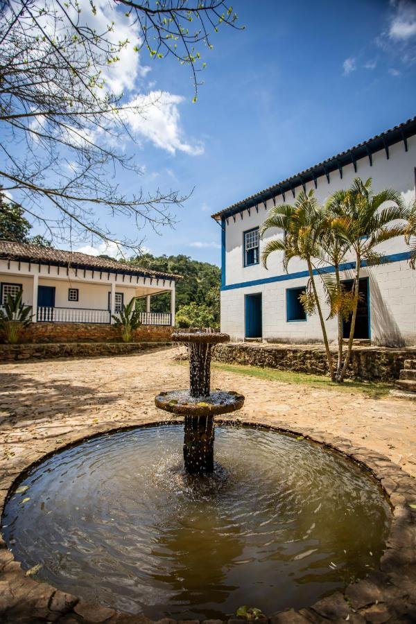 Uma fonte de pedra no centro de um pátio no Hotel Fazenda Paciência com dois edifícios brancos e azuis e palmeiras ao fundo sob um céu azul.