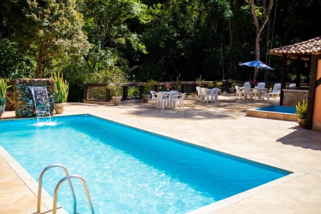 Área de piscina externa com piscina retangular, cascata, espreguiçadeiras brancas, mesas com guarda-sóis e muito verde ao redor no Hotel Fazenda Pirapetinga, um dos hotéis fazenda em Minas Gerais.