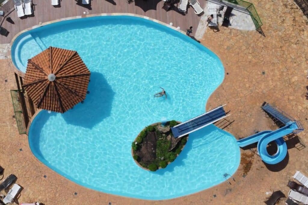 A imagem, vista de cima, mostra uma piscina em formato orgânico com água azul cristalina. No centro da piscina, há uma pequena ilha decorada com plantas, e próximo a ela, um guarda-sol grande com telhado de telhas. À direita, há um tobogã azul que desce em espiral até a piscina. Ao redor da piscina, há uma área de descanso com espreguiçadeiras e mesas brancas, além de um chão de pedra que circunda o espaço. Uma pessoa está relaxando sozinha na água, aproveitando o ambiente ensolarado. Esse é o Hotel Fazenda Poços de Caldas, um dos hotéis-fazenda em Minas Gerais.