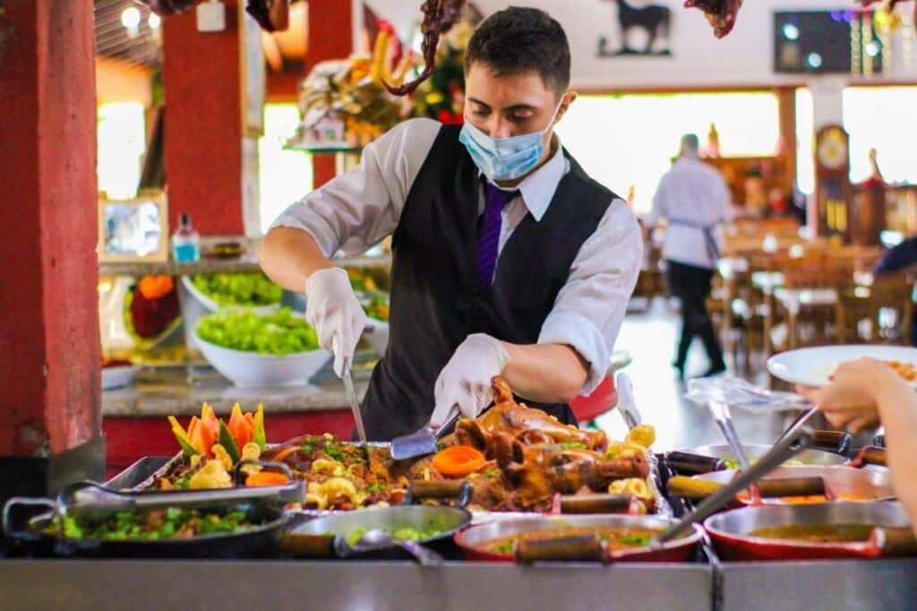 A imagem mostra um atendente de restaurante do Hotel Fazenda Poços de Caldas usando máscara facial e luvas, enquanto serve comida de um buffet variado. Ele está cortando uma carne assada, cercada por diversos pratos coloridos, incluindo legumes, saladas e guarnições. O ambiente parece ser de um restaurante de grande porte, com uma decoração rústica, e ao fundo, há mesas dispostas e outros clientes sendo atendidos. A apresentação dos alimentos é cuidadosamente montada, com detalhes de legumes esculpidos em formato decorativo, realçando o ambiente gastronômico.