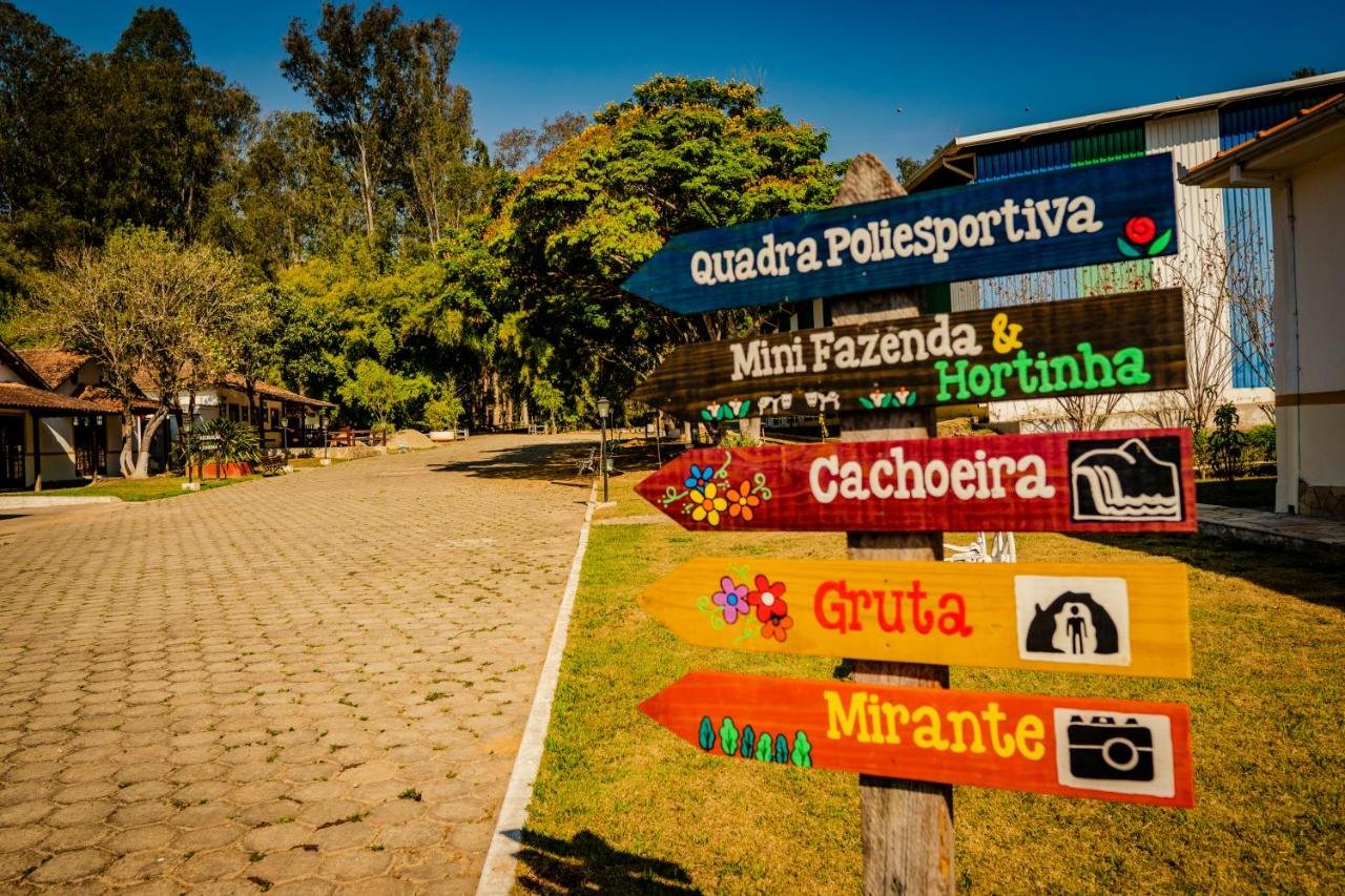 A imagem mostra uma placa de madeira colorida com várias setas apontando para diferentes atrações de um local turístico. As setas indicam direções para uma quadra poliesportiva, uma mini fazenda e hortinha, uma cachoeira, uma gruta e um mirante. As palavras são pintadas de forma decorativa, com ícones ilustrativos ao lado de cada atração, como uma câmera para o mirante e uma caverna para a gruta. Ao fundo, há uma estrada de paralelepípedos ladeada por árvores e construções rústicas, em um cenário de campo ensolarado. Esse é o Hotel Fazenda Ramon, um dos hotéis-fazenda em Minas Gerais.