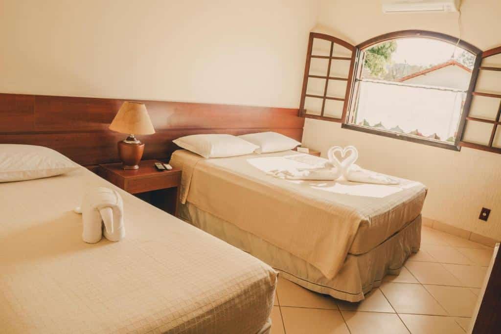 A imagem mostra um quarto do Hotel Fazenda Ramon com uma cama de casal e uma cama de solteiro, ambas cobertas com lençóis brancos. Sobre a cama de casal, há toalhas dobradas em formato de cisnes formando um coração, e sobre a cama de solteiro, uma toalha dobrada em forma de elefante. Entre as camas, há um criado-mudo de madeira com um abajur. O quarto tem uma janela com armação de madeira e vidros abertos, permitindo a entrada de luz natural. O ambiente é simples, acolhedor e bem iluminado, com uma decoração minimalista.