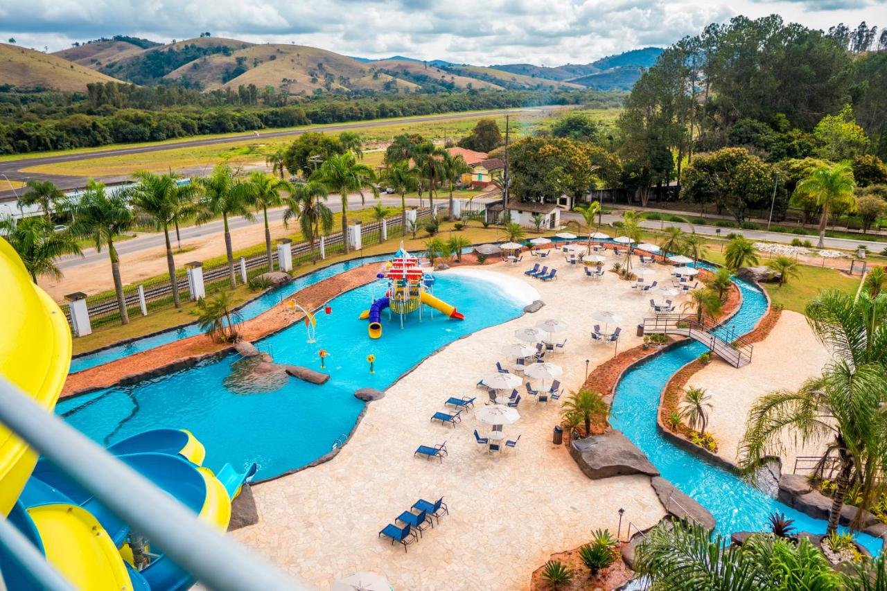 A imagem mostra uma vista aérea de um parque aquático com várias piscinas e áreas de lazer no Hotel Fazenda Ramon. No centro, há uma piscina infantil com um playground aquático colorido, tobogãs e jatos de água. Ao redor, espreguiçadeiras e mesas com guarda-sóis estão dispostas em uma área pavimentada. Há um rio artificial que serpenteia pela área, rodeado de plantas e pontes decorativas. No fundo, vê-se uma paisagem montanhosa verde, com palmeiras ao longo de uma estrada próxima, completando o cenário relaxante e familiar.