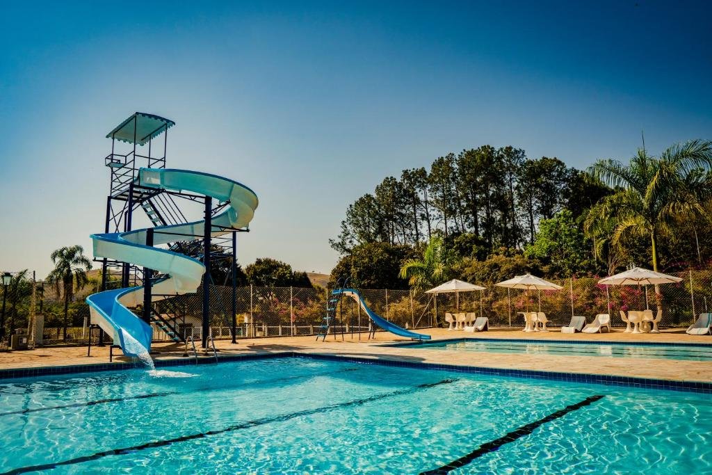 A imagem mostra uma piscina em um parque aquático, com um grande toboágua em espiral de cor azul claro. O toboágua começa em uma estrutura alta de metal e desce em curvas até a piscina, onde a água está clara e convidativa. Ao lado, há uma pequena rampa aquática azul que termina diretamente na piscina. Em volta da piscina, estão dispostas espreguiçadeiras e mesas brancas sob guarda-sóis, proporcionando áreas de descanso. Ao fundo, há uma cerca e árvores verdes, com um céu claro e azul, criando um ambiente agradável e ensolarado. Esse é o Hotel Fazenda Ramon, um dos hotéis-fazenda em Minas Gerais.