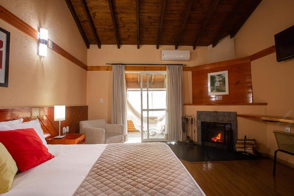 Quarto do Hotel Fazenda Rota dos Cânions com cama de casal, lareira, poltrona e vista para um pátio e rede através de portas de vidro deslizantes.