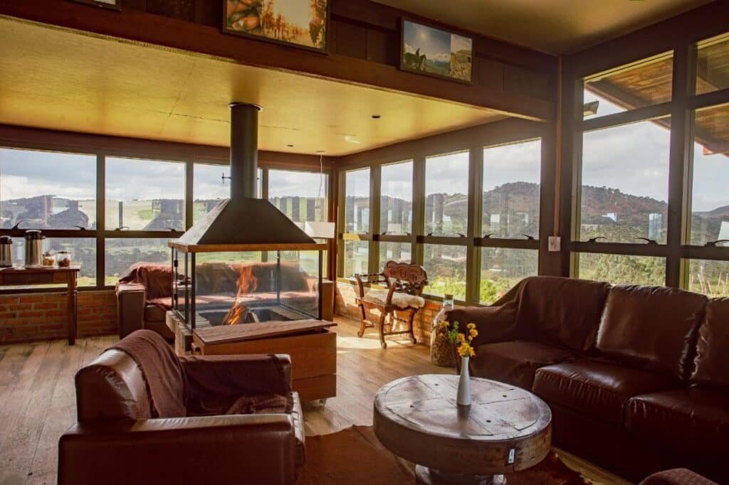 Uma sala de estar aconchegante no Hotel Fazenda Rota dos Cânions com grandes janelas, lareira central, sofás de couro, uma mesa redonda de madeira com flores e vista para as montanhas.