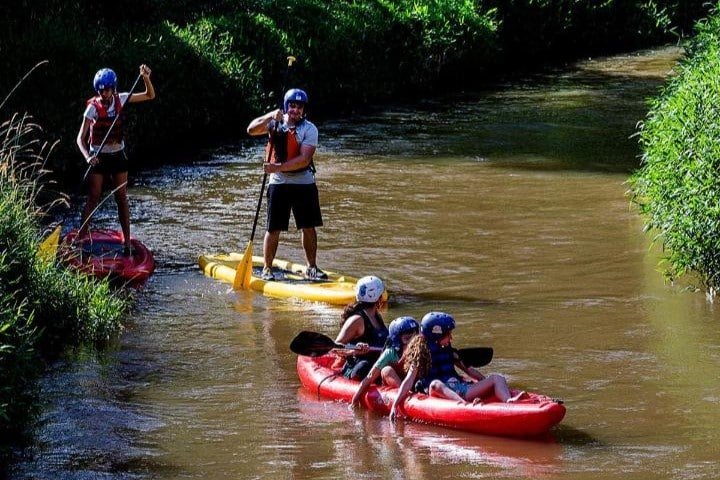 Três pessoas praticando paddleboard e três pessoas praticando caiaque em um rio estreito e lamacento cercado por vegetação densa no Hotel Fazenda Terra dos Sonhos. Todos os participantes usam capacetes.