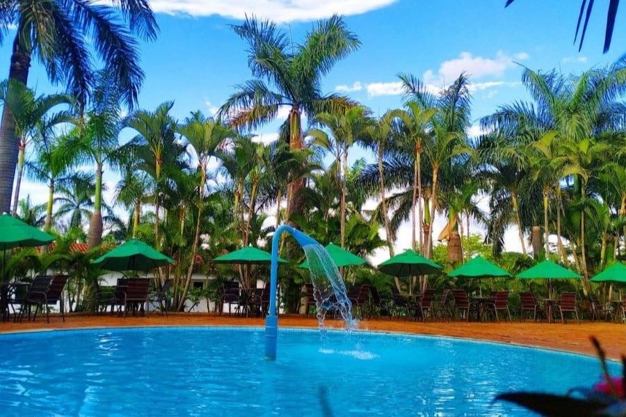 A imagem mostra uma piscina circular com uma fonte de água central, localizada em um ambiente cercado por muitas palmeiras altas e verdes. Ao redor da piscina, há várias mesas e cadeiras com guarda-sóis verdes, criando uma área confortável para descanso à sombra. O céu está azul com algumas nuvens, contribuindo para o clima tropical e relaxante do local. A vegetação exuberante e a organização da área de lazer oferecem um ambiente convidativo para relaxar e aproveitar o dia ao ar livre. Esse é o Hotel Fazenda Vale Amanhecer, um dos hotéis-fazenda em Minas Gerais.