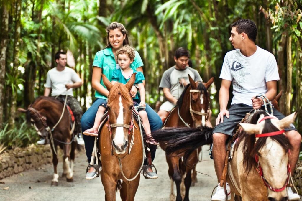 Um grupo de pessoas está cavalgando por uma trilha florestal no Hotel Fazenda Vale das Pedras. Uma mulher tem uma criança pequena com ela em um cavalo.