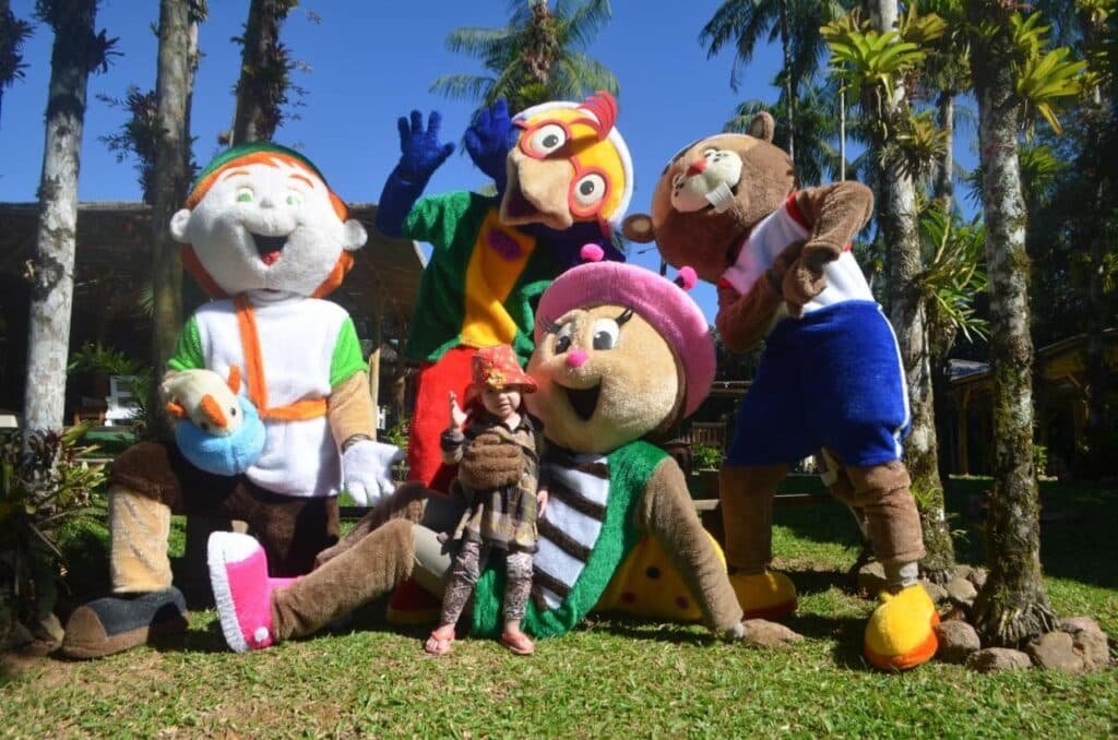 Uma criança posa com quatro pessoas em fantasias coloridas de animais de desenho animado em uma área gramada com palmeiras ao fundo no Hotel Fazenda Vale das Pedras, um dos hotéis fazenda em Santa Catarina.