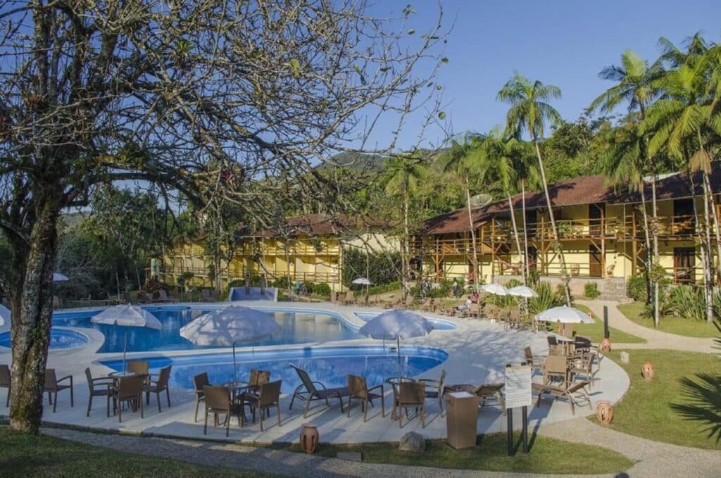 Uma área de piscina com mesas, cadeiras e guarda-sóis em frente a edifícios cercados por árvores e vegetação em um dia claro. Esse é o Hotel Fazenda Vale das Pedras, um dos hotéis fazenda em Santa Catarina.