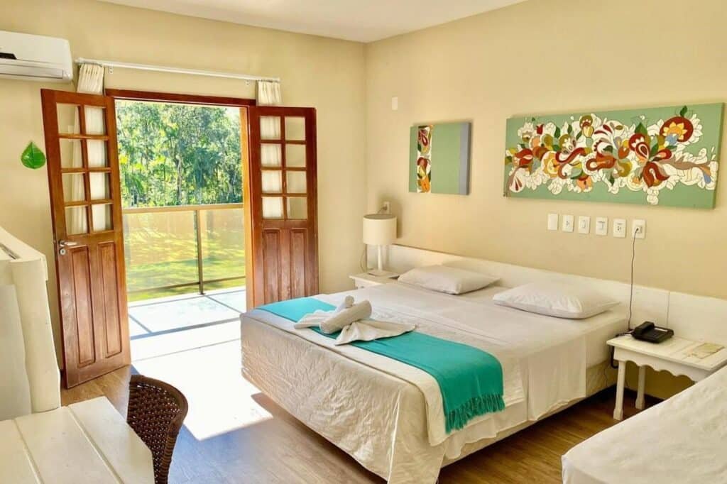 Um quarto iluminado pelo sol no Hotel Fazenda Vale das Pedras com uma cama de casal, obras de arte e portas francesas de madeira que se abrem para uma varanda com vista para a vegetação verde.