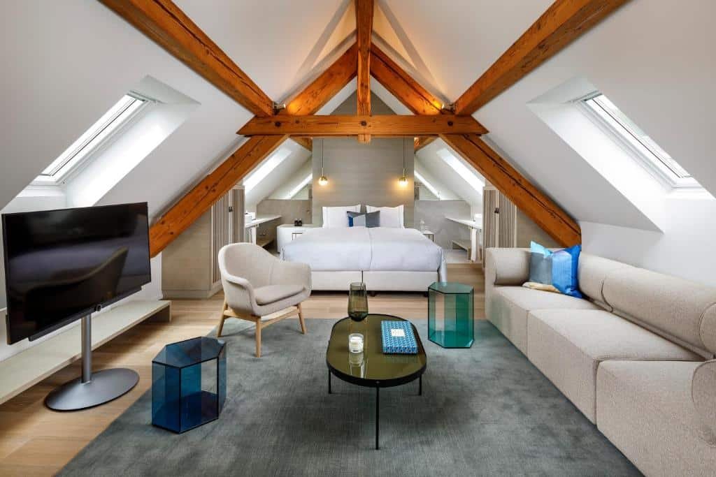 Interior do loft do Hotel Felix, de 58 m², com sala de estar com sofá cinza, TV, mesa redonda no centro, poltrona cinza e cama de casal branca com algumas almofadas e travesseiros em cima
