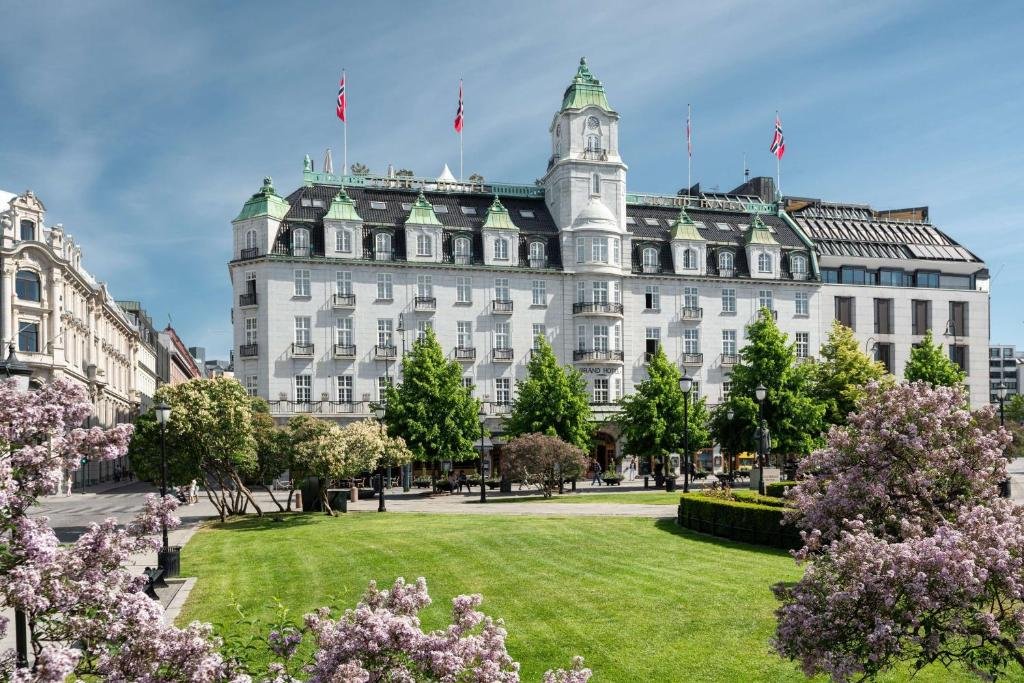 Frente do Grand Hotel edificio em tom branco com telhados cinzas e um jardim com árvores e grama verde a frente dele. Representa hotéis em Oslo