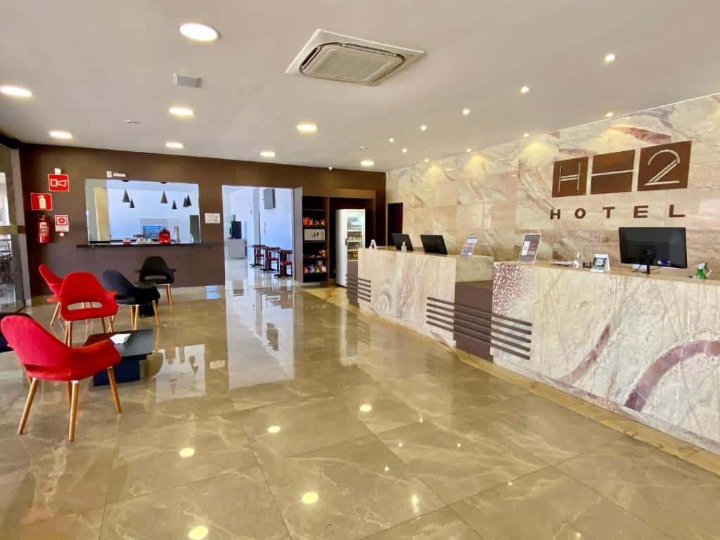 hall de entrada do Hotel H2 Congonhas com uma bancada de pedra extensa, onde é possível ver três monitores de computador pretos. Ao fundo há uma ampla passagem sem porta e uma janela ao lado mostrando o refeitório