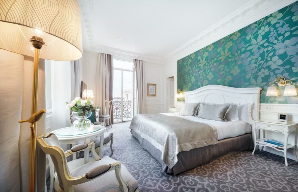Quarto do Hôtel Hermitage Monte-Carlo. Uma cama de casal no lado direito com um criado mudo de cada lado. De frente para a cama, uma poltrona com abajur e uma mesa redonda. No fundo do quarto a sacada. Foto para ilustrar post sobre Hotéis em Mônaco.