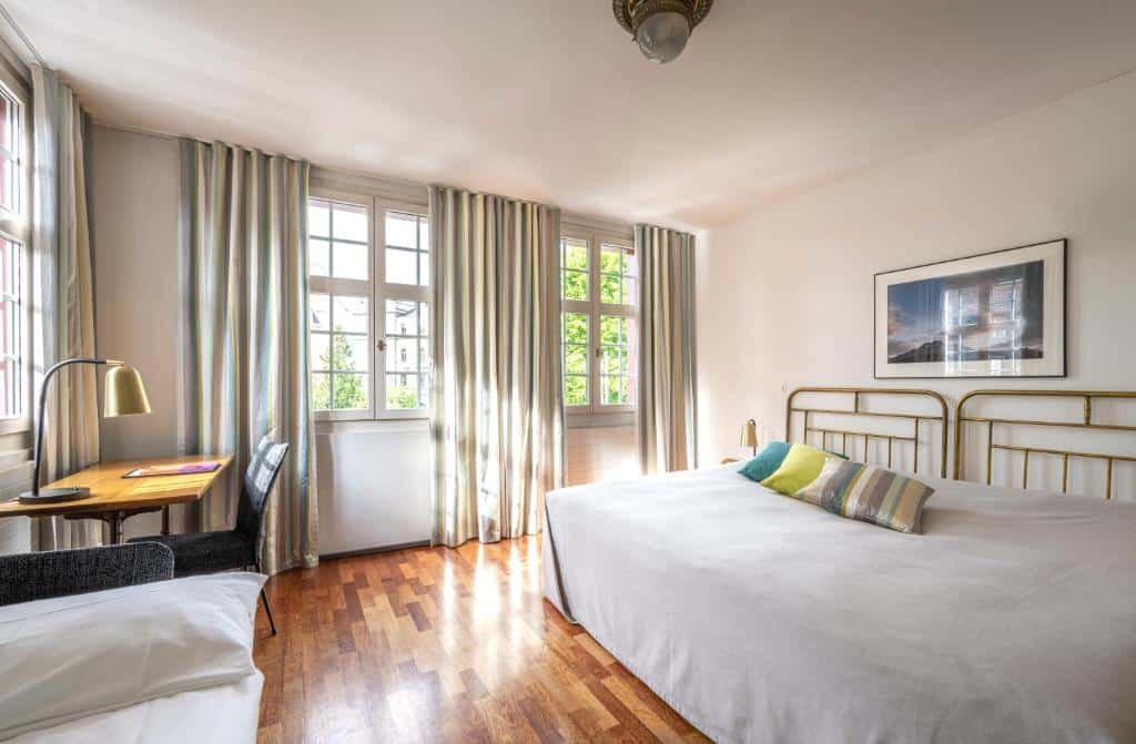 Foto do Hotel Hofgarten Luzern, para representar o post de hotéis em Lucerna. O ambiente é bem espaçoso. Na direita vemos uma extensa cama de casal. Na esquerda há visão parcial de um sofá no canto inferior da foto e de uma escrivaninha ao seu lado. Há janelas espalhadas pelo quarto.