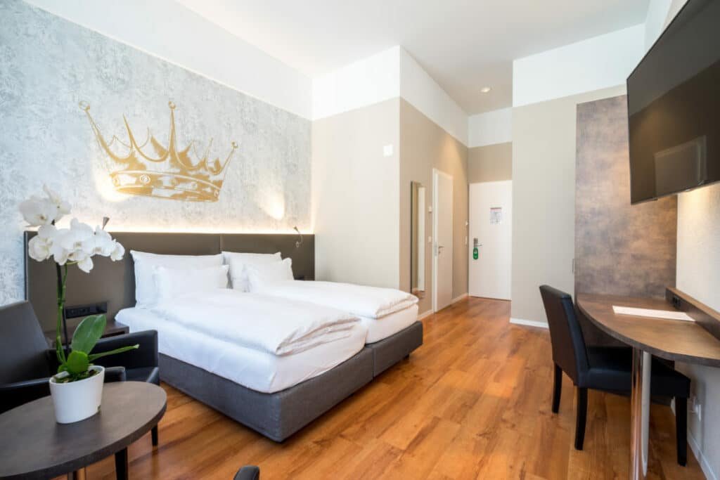 Foto do Altstdat Hotel Krone Luzern, para representar o post de hotéis em Lucerna. O quarto possui uma cama de casal box, que está encostada na parede na esquerda. Atrás, possui a logo de uma coroa, marca do hotel. No canto inferior esquerdo é possível ver uma pequena mesa redonda com vaso de flor em cima. Já no lado direito, na frente da cama, encostada na parede, está uma mesa com uma cadeira. E há uma televisão em cima fixa na parede.