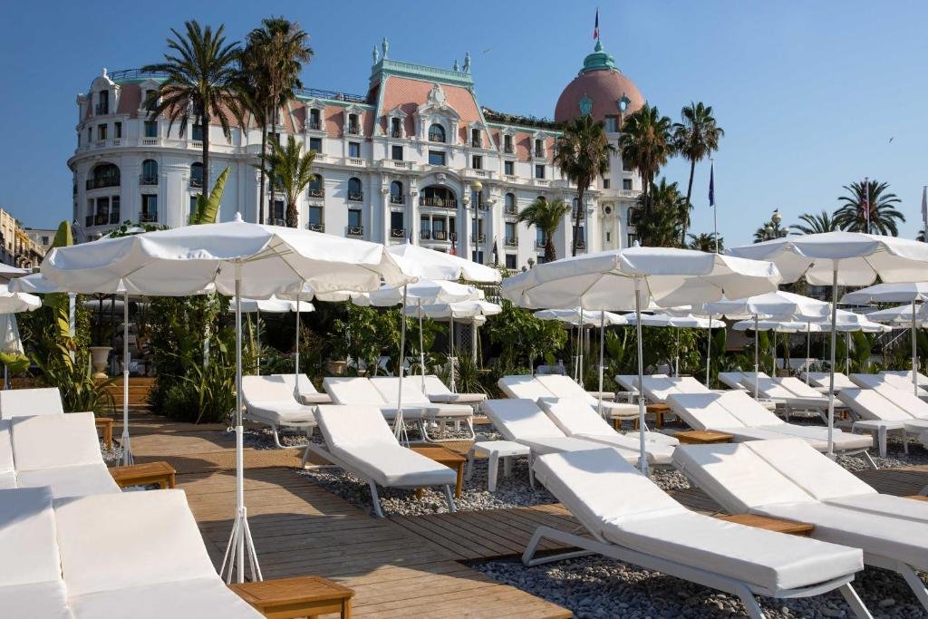 Praia em frente ao Hotel Le Negresco. Na parte da frente há diversas espreguiçadeiras com guarda-sóis e no fundo é possível ver o hotel.