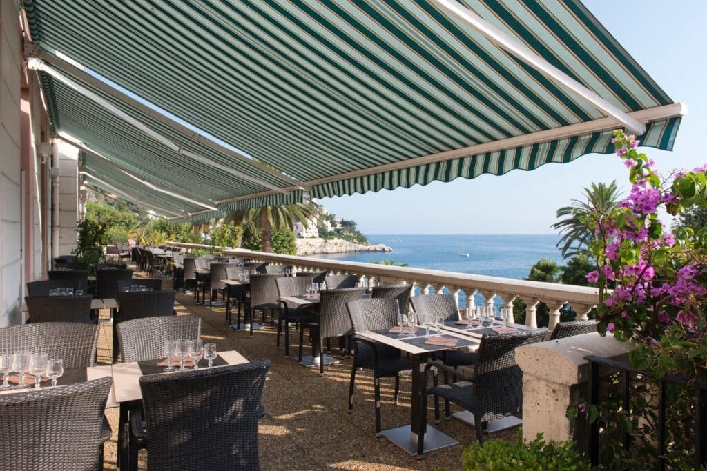 Área externa de refeições do Hotel Le Saint Paul. Diversas mesas e cadeiras estão enfileiradas em um terraço coberto logo ao lado do mar.