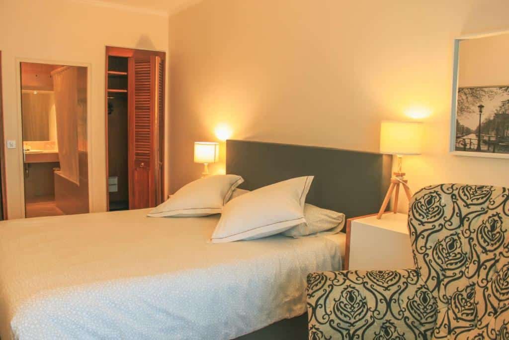 Foto de um quarto no Hotel Les Arcades, para ilustrar o post de hotéis em Genebra. A cama é de casal e está no centro da foto, temos uma visão lateral dela. Em seus dois lados há uma mesa de escrivaninha com abajur em cima. No canto inferior direito, há uma poltrona. Já na esquerda, vemos o closet de madeira e uma porta para o banheiro.