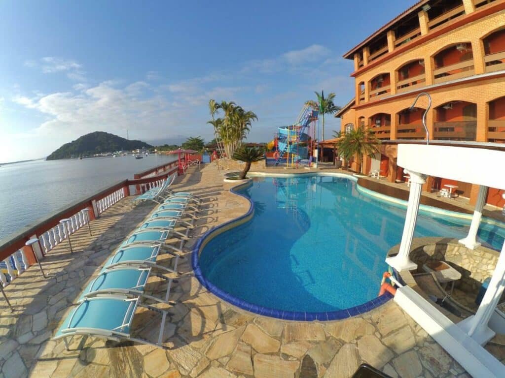Área de lazer do Hotel Marazul. Uma pisicna está em primeiro plano e, ao seu lado, estão váris espreguiçadeiras. Ao fundo é possíver ver um grande escorregador. Ao lado de tudo isso está o mar.