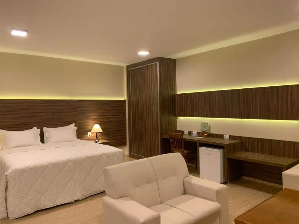 quarto do Hotel Mirante São Brás em São Brás do Suaçuí, próximo a Congonhas, com uma cama de casal ao centro, um pequeno sofá de dois lugares, branco, em frente, uma mesa de madeira com cadeira e um frigobar embaixo no lado direito da cama.