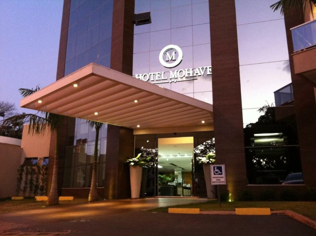 Faixada espelhada do hotel com porta de entrada, luzes, algumas árvores e uma placa de estacionamento para pessoas com deficiência.
