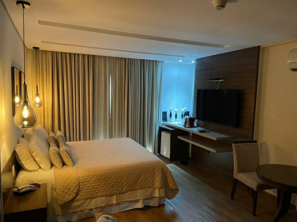 Quarto com uma cama de casal e luminárias penduradas no teto. É possível ver um frigobar, uma televisão em frente a cama e uma mesa com cadeira no canto. Do outro lado há uma janela com cortinas. Foto para ilustrar post sobre hotéis em Campo Grande.