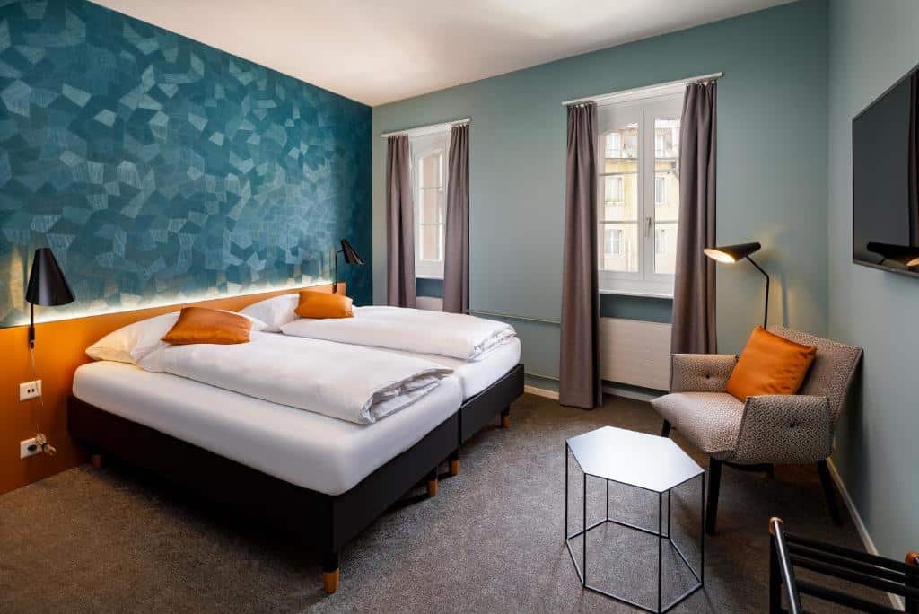 Quarto do Hotel Münzgasse. A cama box de casal está encostada na parede à esquerda. Do lado direito da cama, na parede no fundo, há duas janelas. No lado direito do quarto, de frente com a cama, há uma TV na parede, uma poltrona, abajur e uma mesa de centro. No canto inferior direito da foto é possível identificar um suporte para passar roupas.