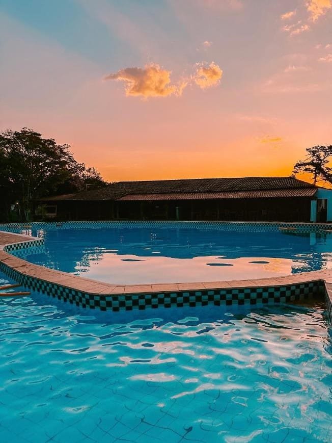 Uma piscina com água azul clara durante o pôr do sol, com um edifício e árvores ao fundo sob um céu colorido. O local fica no Hotel Pontal de Tiradentes, e faz parte da lista de hotéis fazenda em Minas Gerais.