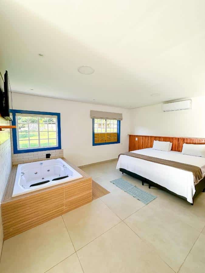 Um quarto do Hotel Pontal de Tiradentes dispõe de uma cama de casal, uma televisão montada na parede, uma jacuzzi e duas janelas com molduras azuis e persianas bege.