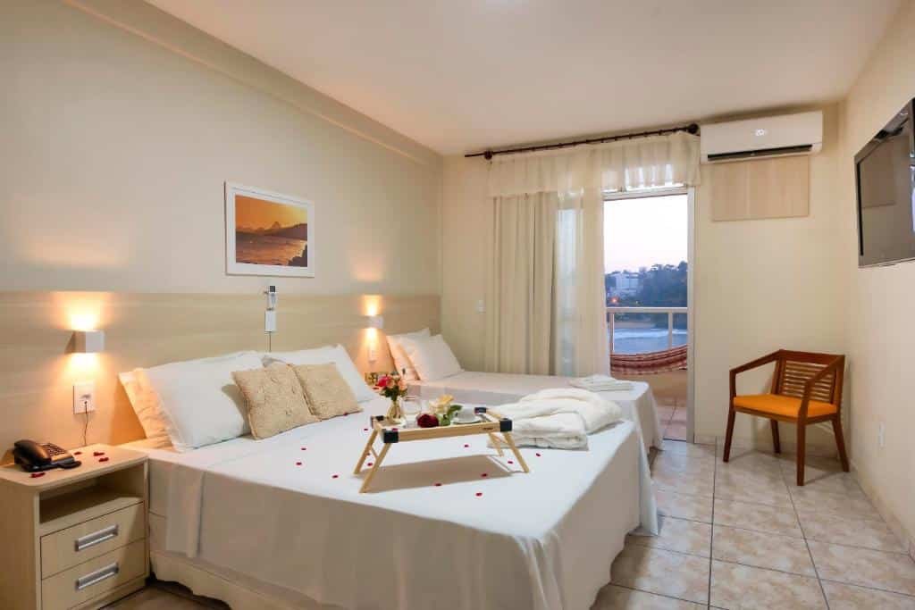 Quarto do Hotel Portal da Lua. Uma cômoda com um telefone do lado esquerdo, e uma cama de casal e de solteiro. De frente para a cama uma televisão e uma cadeira. No fundo, a sacada do quarto com uma rede e vista do mar. Em cima da cama, uma mesinha de madeira com flor, copo e xícara.