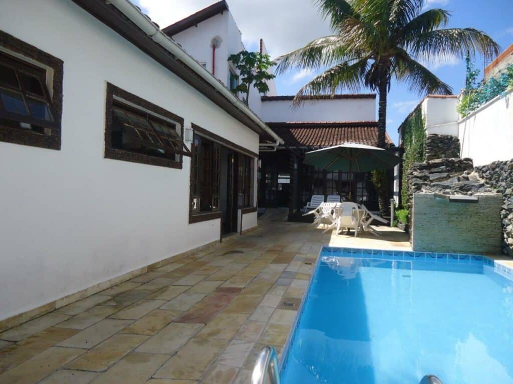 Piscina do Hotel Pousada da Néia. Ela é retangular e está no canto direito, em uma área mais estreita. Ao fundo estão uma mesa, cadeiras e guarda-sol.