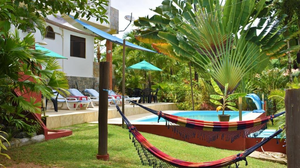 Área externa do Hotel Pousada Rancho Fundo. Duas redes na frente, atrás uma piscina e cadeiras de tomar sol com guarda-sóis, mesas e cadeiras, no fundo várias árvores.