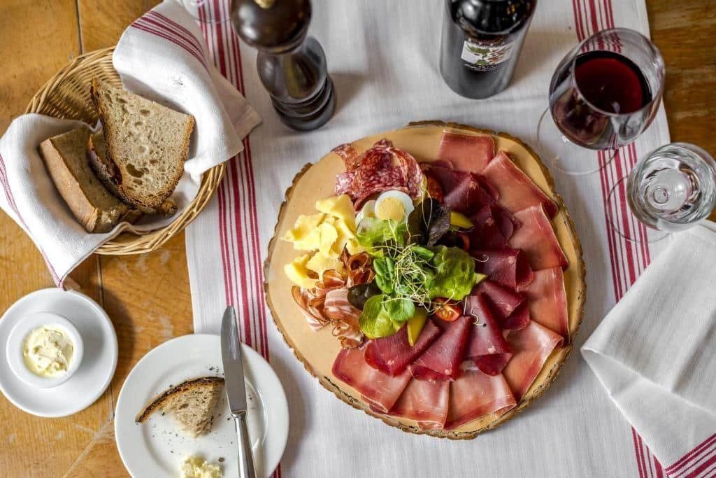 Uma foto de uma refeição sendo realizada no Hotel Rebstock. No centro há um prato mais largo com petiscos e frios que incluem tiras de salame, ovos, queijos, verduras, bacon, etc. Na direita, há um guardanapo e uma taça com suco de uva. Na esquerda, uma cesta com pães integrais e um prato com um pouco de manteiga.