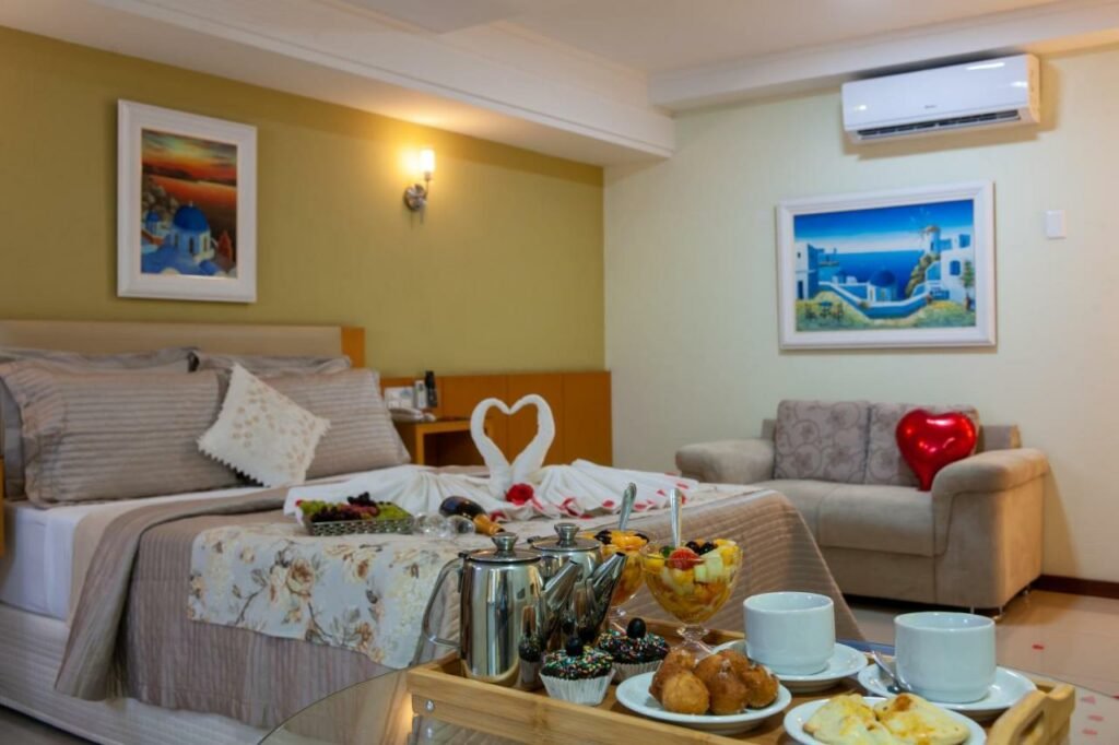 Quarto no Hotel Santorini com uma cama de casal, uma mesa repleta de comidas e bebidas, e um ambiente romântico com tudo em formato de coração