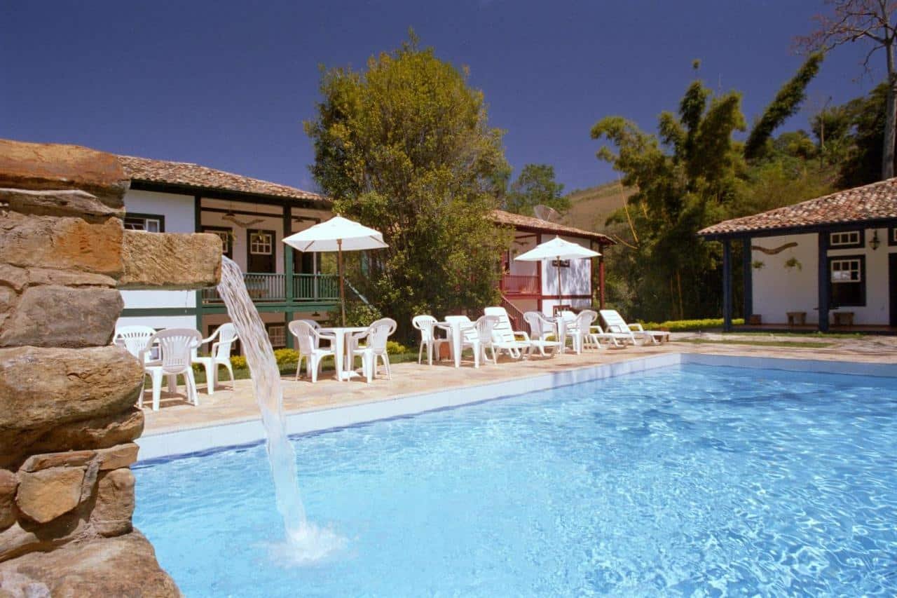 Área de piscina ao ar livre com cadeiras de jardim, guarda-sóis e edifícios rústicos ao fundo, cercada por vegetação e céu azul. O local fica no Hotel Solar dos Montes, um dos hotéis fazenda em Minas Gerais.