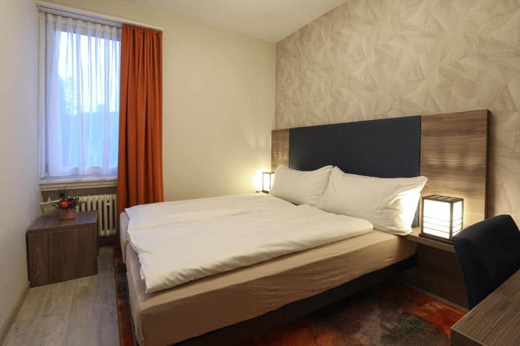 Foto do Hotel Spatz, para representar o post de hotéis em Lucerna. A cama é box de casal e está encostada na parede da direita. Assim, temos uma visão lateral sua. No canto inferior direito da foto temos uma visão parcial de uma mesa com cadeira. No fundo do quarto, na esquerda, está uma janela com cortinas vermelhas. O ambiente não é muito espaçoso, mas também não é extremamente apertado.