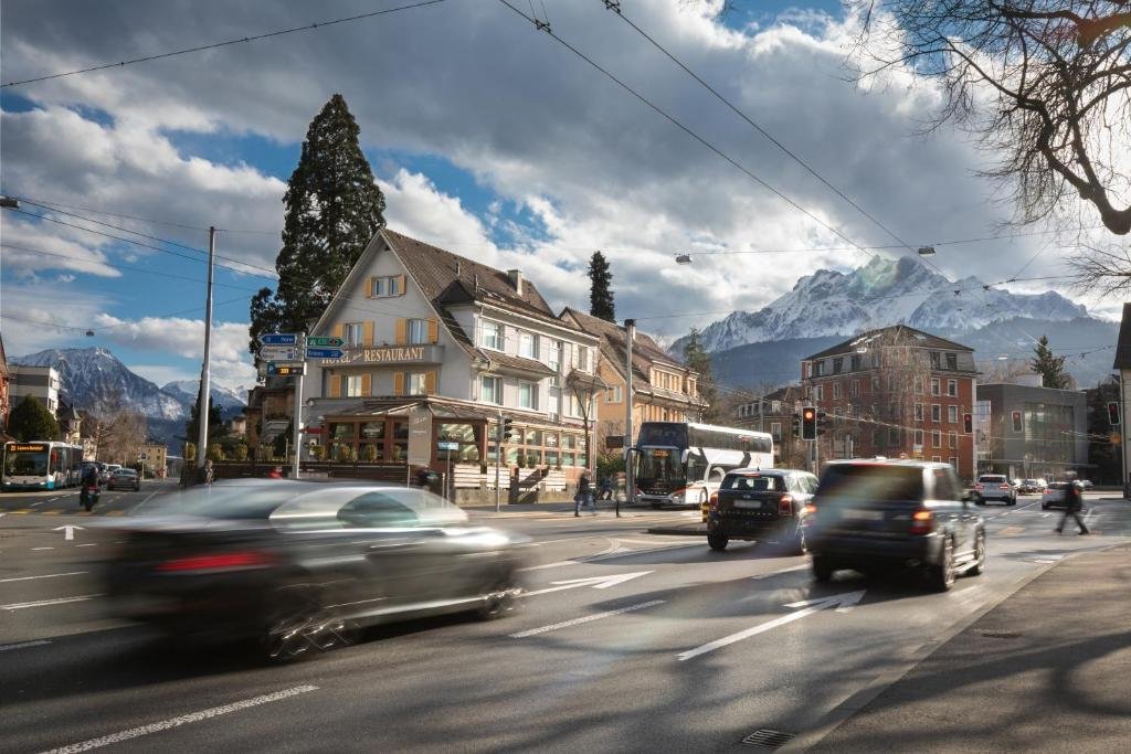 Foto da rua em que está localizado o Hotel Spatz. Carros estão passando pela avenida, e do outro lado está o hotel, perto de outros prédios. Ao fundo vemos montanhas e até mesmo teleféricos passando. A foto foi tirada de dia mas há nuvens carregadas no céu.