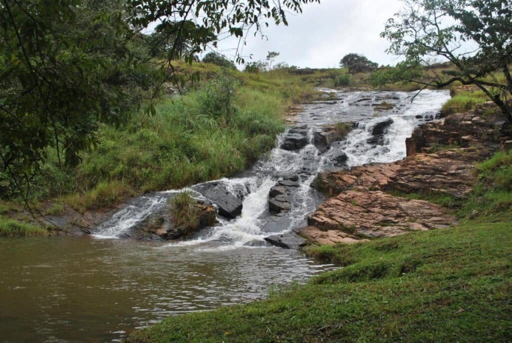 A imagem mostra uma bela cachoeira fluindo por rochas escuras em uma área cercada por vegetação densa e gramada no Hotel Termópolis. A água desce em várias quedas pequenas, formando um curso d