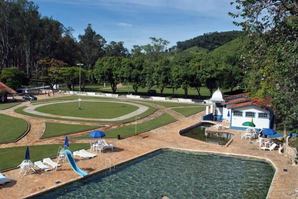 A imagem mostra uma ampla área externa em um local de lazer com uma piscina grande no primeiro plano. Ao redor da piscina, há espreguiçadeiras brancas e guarda-sóis azuis, além de um pequeno toboágua. O piso ao redor da piscina é de pedra, e há caminhos que conduzem a um prédio com telhado de cerâmica à direita. O cenário é cercado por árvores e uma extensa área gramada com um caminho circular pavimentado no centro. Ao fundo, colinas verdes e árvores completam o cenário tranquilo e convidativo. Esse é o Hotel Termópolis, um dos hotéis-fazenda em Minas Gerais.