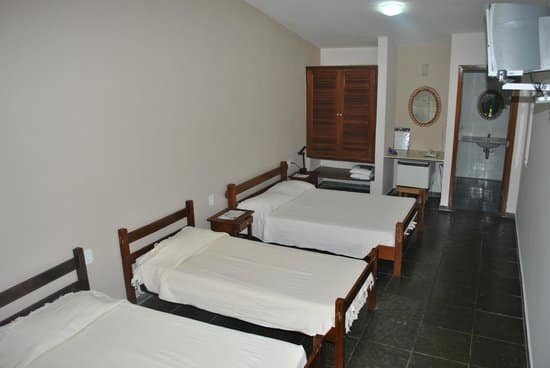 A imagem mostra um quarto simples do Hotel Termópolis com três camas de solteiro dispostas em linha, todas cobertas com lençóis brancos. O quarto tem piso escuro e uma parede pintada em tom claro. No fundo do quarto, há uma janela de madeira fechada e, ao lado, uma pequena bancada com uma pia, toalhas dobradas e um espelho redondo na parede. Ao fundo, há uma porta que parece levar ao banheiro. O ambiente é básico e funcional, com um estilo limpo e prático.