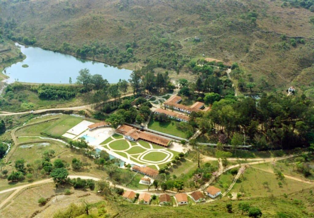 A imagem mostra uma vista aérea de uma fazenda ou hotel rural cercado por colinas e vegetação. No centro, há várias construções com telhados de telha vermelha, incluindo uma área com piscina e jardins bem cuidados com caminhos desenhados. À esquerda, pode-se ver um lago cercado por árvores, e estradas de terra conectam as áreas da propriedade. O ambiente natural ao redor é vasto, com vegetação esparsa e áreas montanhosas, transmitindo tranquilidade e isolamento em meio à natureza. Esse é o Hotel Termópolis, um dos hotéis-fazenda em Minas Gerais.