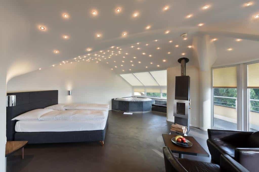 Quarto do Hotel Uto Kulm, com decoração clean, várias luzes no teto simulando estrelas brilhando, além de cama de casal grande, hidro, sofá com mesa pequena quadrada no meio, lareira e janelas mostrando a natureza verde em volta
