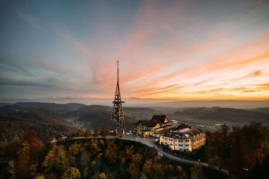 Região onde fica o Hotel Uto Kulm, em Zurique, em um fim de tarde com o céu laranja, com muita natureza em volta, e o prédio do local no meio com uma torre ao lado