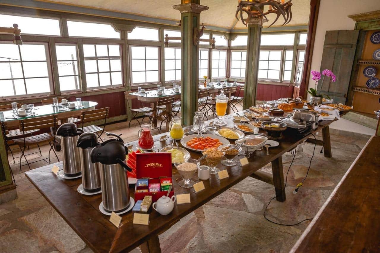 A imagem mostra uma mesa grande e rústica de madeira com um farto buffet de café da manhã do Hotel Vila Relicário. Entre os itens, há jarras com sucos de frutas, potes com cereais, tigelas de frutas como mamão e melão, pães, bolos e opções de chá. Ao lado da mesa, há cafeteiras térmicas e um expositor de chás da marca Twinings. Ao fundo, mesas de vidro com cadeiras de madeira estão preparadas para os convidados. O ambiente é iluminado naturalmente por amplas janelas, e a decoração é aconchegante, com detalhes em madeira e tons acolhedores.