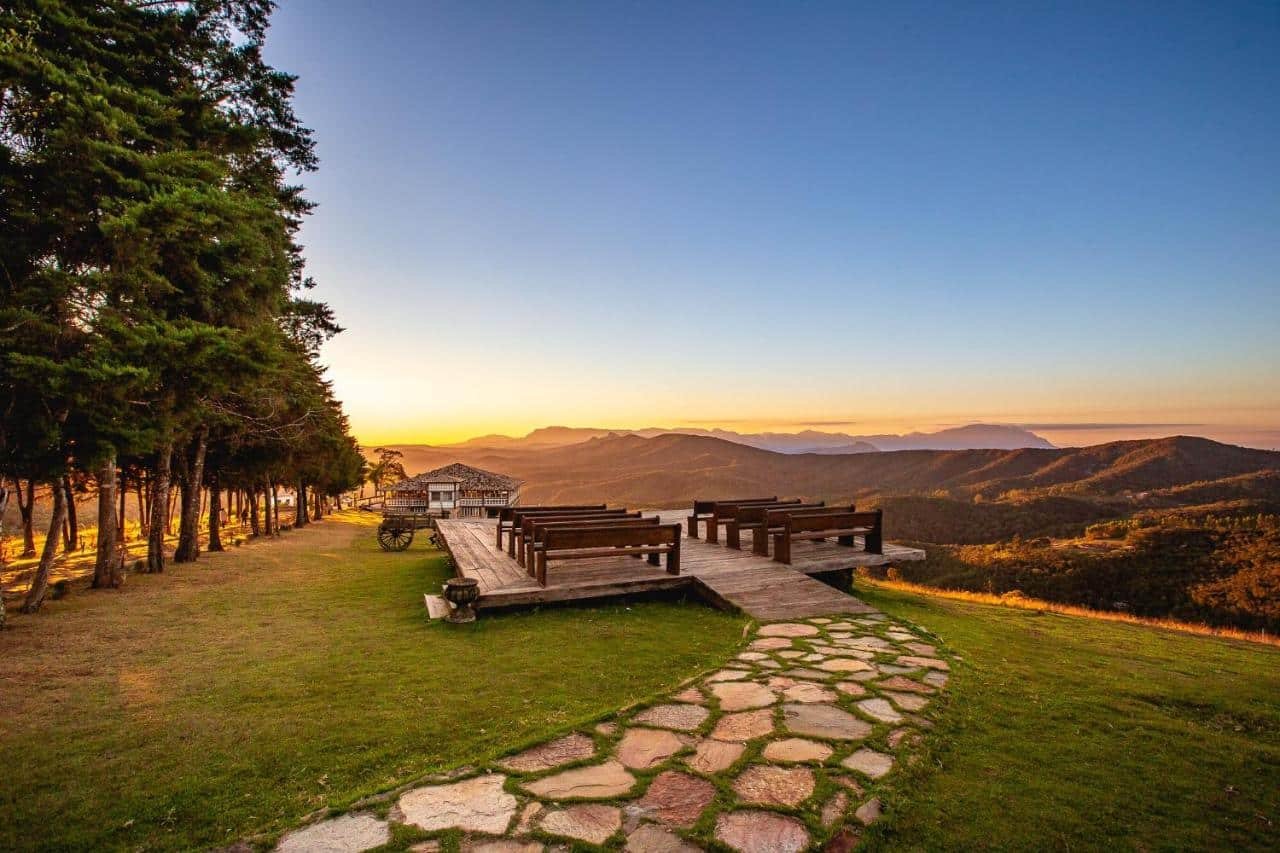A imagem mostra uma área ao ar livre com um deck de madeira elevado, com bancos de madeira dispostos em fileiras, voltados para uma vista deslumbrante das montanhas ao pôr do sol. O céu está claro, com tons dourados e laranja, iluminando suavemente o horizonte. À esquerda, há uma fileira de árvores altas, e o gramado verde se estende até o deck, com um caminho de pedras que leva até a área principal. Ao fundo, também é possível ver uma pequena casa rústica de telhado de telhas. A paisagem é tranquila e oferece uma vista panorâmica das montanhas. Esse é o Hotel Vila Relicário, um dos hotéis-fazenda em Minas Gerais.