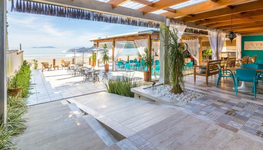 Área externa do Hotel Village Enseada, um hall extenso coberto e cheio de mesas e cadeiras que ficam já em direção da praia, com a areia logo em seguida, tudo decorado de um jeito rústico